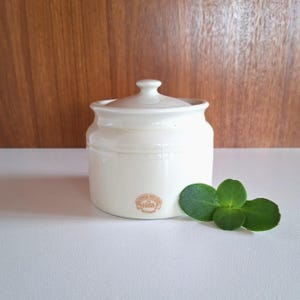 May include: A cream-coloured ceramic jar with a matching lid and a rounded knob handle. The front features a small circular stamp with the text Bendigo Pottery 1858 Australia. A small green succulent plant rests beside the base of the jar.