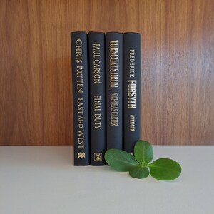 Puede incluir: Cinco libros de tapa dura negros con letras doradas en el lomo. Los libros se titulan "East and West" de Chris Patten, "Final Duty" de Paul Carson, "Turncoat's Drum" de Nicholas Carter y "Avenger" de Frederick Forsyth. Una hoja verde est&aacute; delante de los libros.
