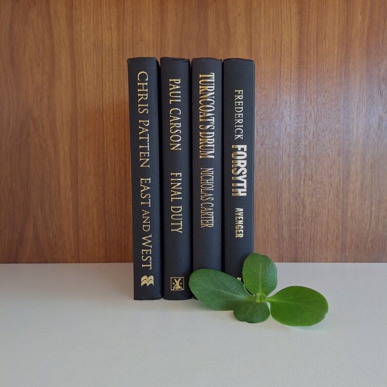 Puede incluir: Cinco libros de tapa dura negros con letras doradas en el lomo. Los libros se titulan "East and West" de Chris Patten, "Final Duty" de Paul Carson, "Turncoat's Drum" de Nicholas Carter y "Avenger" de Frederick Forsyth. Una hoja verde est&aacute; delante de los libros.