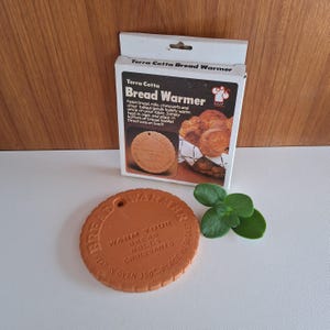 May include: A terra cotta bread warmer with a round shape and the text "Warm your bread, rolls, croissants" is displayed. The warmer is next to its original box, which has the words "Terra Cotta Bread Warmer" printed on it.