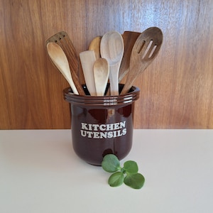 May include: A dark brown ceramic kitchen utensil holder with the words "KITCHEN UTENSILS" in white. The holder contains various wooden spoons and spatulas. A small green plant is in front. The background is a wooden surface.
