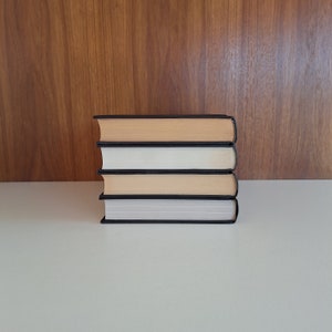 Puede incluir: Una pila de cuatro libros con cubiertas negras y p&aacute;ginas de color crema. Los libros est&aacute;n apilados sobre una superficie blanca.