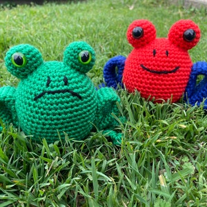 May include: Two crocheted frog plushies, one green with a sad expression and one red with a happy expression. The green frog is sitting on the grass, while the red frog is sitting on the grass with its blue arms outstretched.