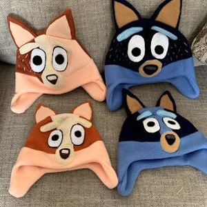 May include: Four fleece hats featuring cartoon dog character faces. Two hats are light orange and brown with pointed ears, and two hats are dark blue and light blue with pointed ears. Each hat has large white eyes, a nose, and ear flaps.