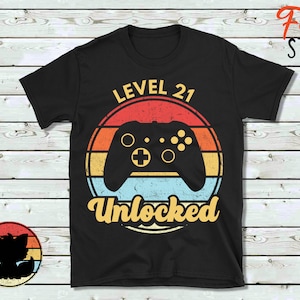 May include: Black t-shirt with a retro-style graphic of a video game controller and the text "Level 21 Unlocked".