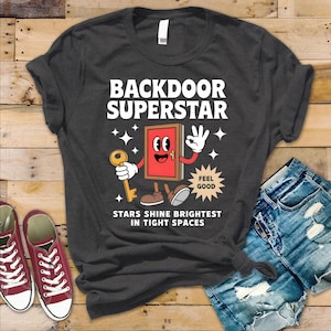 May include: A dark grey t-shirt with a white graphic of a red door with a keyhole and a smiling face. The text "Backdoor Superstar" is above the door and "Stars shine brightest in tight spaces" is below the door. A speech bubble above the door says "Feel good".