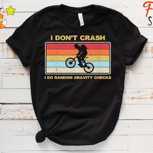 以下が含まれることがあります： 黒いTシャツ。レトロなデザインで、マウンテンバイクに乗ったサイクリストが描かれています。「I DON'T CRASH I DO RANDOM GRAVITY CHECKS」というテキストが、赤、オレンジ、黄色、青、白の水平ストライプが入った長方形の中にあります。
