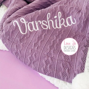 May include: A purple cable knit blanket with the name "Varshika" embroidered in white. The blanket is folded and has a white fluffy edge. The logo "Love Jo Designs Embroidered Clothing" is visible in the bottom right corner.