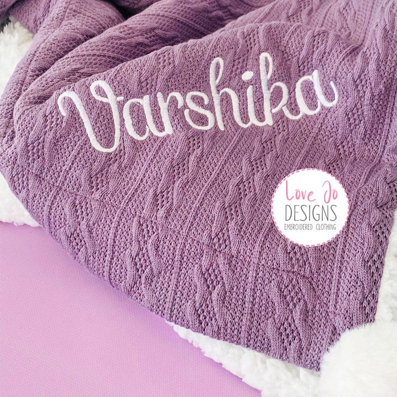 May include: A purple cable knit blanket with the name "Varshika" embroidered in white. The blanket is folded and has a white fluffy edge. The logo "Love Jo Designs Embroidered Clothing" is visible in the bottom right corner.