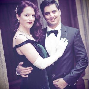 May include: A couple in formal attire, the woman wearing a green velvet dress with white gloves and the man wearing a black tuxedo with a bow tie. The couple is standing in front of a stone wall.