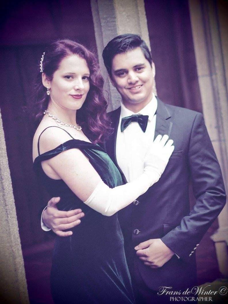 May include: A couple in formal attire, the woman wearing a green velvet dress with white gloves and the man wearing a black tuxedo with a bow tie. The couple is standing in front of a stone wall.
