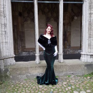 May include: A woman in a dark green velvet gown with a black fur shawl and white gloves stands in a stone archway.
