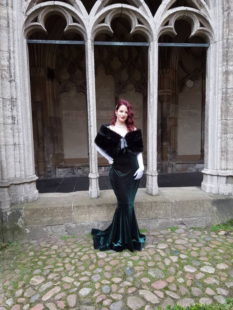 May include: A woman in a dark green velvet gown with a black fur shawl and white gloves stands in a stone archway.