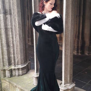 May include: A woman in a long, dark green velvet gown with a black fur shawl and white gloves stands in a stone archway.