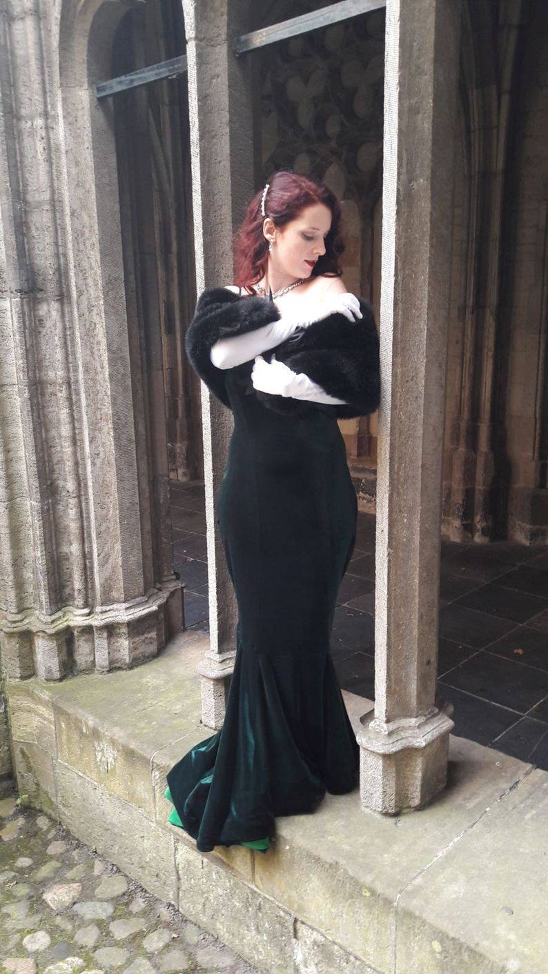 May include: A woman in a long, dark green velvet gown with a black fur shawl and white gloves stands in a stone archway.