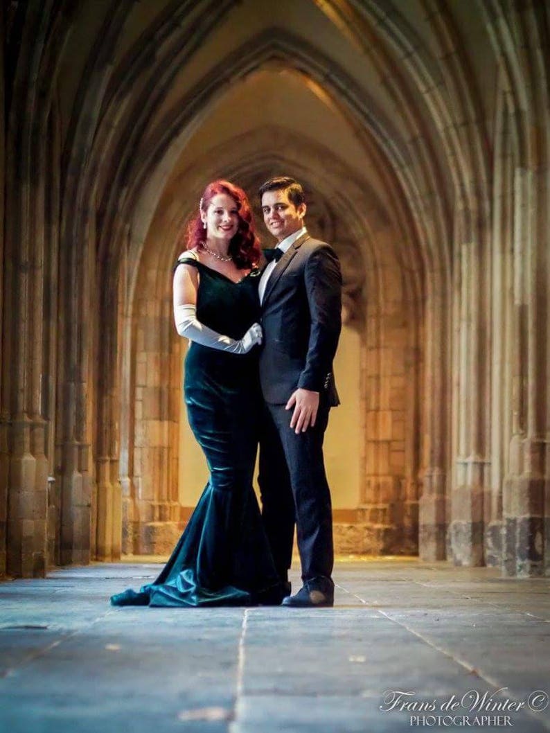 May include: A couple poses for a portrait in a stone archway. The woman is wearing a dark green velvet gown with a white lace overlay and white gloves. The man is wearing a dark suit with a white shirt and a dark tie.