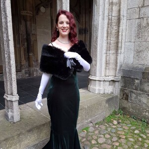 May include: A woman wearing a long, dark green velvet gown with a black fur shawl and white gloves. She is standing in front of a stone building with an arched doorway.