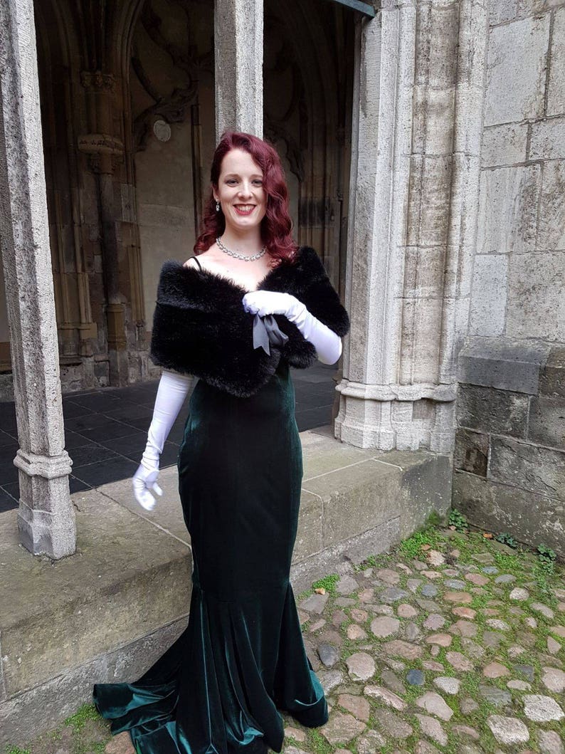 May include: A woman wearing a long, dark green velvet gown with a black fur shawl and white gloves. She is standing in front of a stone building with an arched doorway.
