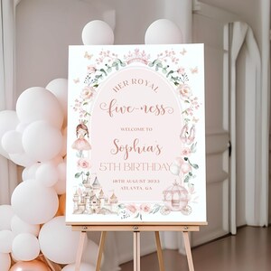 May include: A pink and white welcome sign on a wooden easel. The sign features floral and princess-themed illustrations and text that reads "Her Royal Fiveness" and "Welcome to Sophia's 5th Birthday". White and rose gold balloons are in the background.