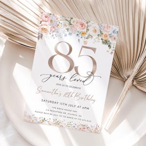 May include: An invitation card for an 85th birthday, with floral decorations. The text reads "85 years loved" and includes event details. The card is set on a white plate with a dried palm leaf.