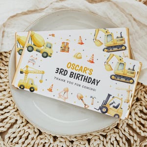 Könnte beinhalten: Zwei Schokoriegel mit Motiven zum Thema Bau. Ein Riegel trägt den Text "OSCAR'S 3RD BIRTHAY" und Bau-Fahrzeuge. Der andere Riegel hat ähnliche Illustrationen. Die Riegel liegen auf einem weißen Teller.