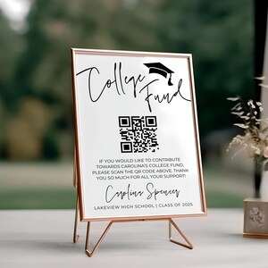 May include: A white sign with a black graduation cap and the text "College Fund" in black script. A black and white QR code is below the text. The sign also includes the text "If you would like to contribute towards Carolina's college fund, please scan the QR code above. Thank you so much for all your support!" and "Caroline Spencer Lakeview High School | Class of 2025". The sign is on a gold metal stand.
