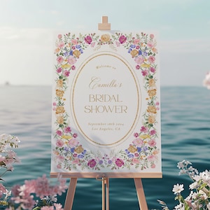 Puede incluir: Un cartel blanco con un borde floral y letras doradas anunciando una despedida de soltera para Camilla. El texto dice "Bienvenida a la despedida de soltera de Camilla el 18 de septiembre de 2023 en Los &Aacute;ngeles, California."