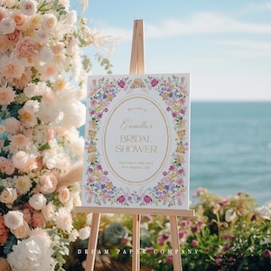 Puede incluir: Un cartel blanco con un borde floral y letras doradas que anuncian "Bienvenido al Bridal Shower de Camilla" con la fecha y la ubicaci&oacute;n. El cartel est&aacute; en un caballete frente a un arco floral.