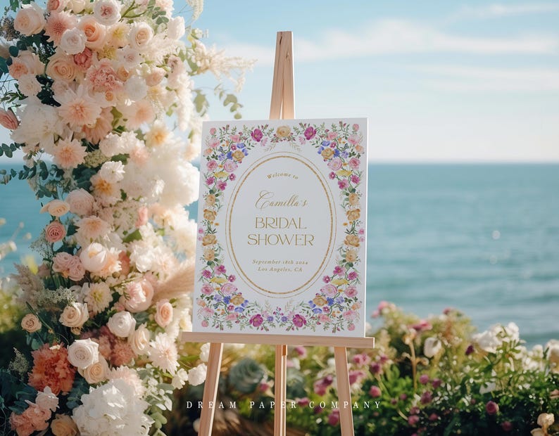 Puede incluir: Un cartel blanco con un borde floral y letras doradas que anuncian "Bienvenido al Bridal Shower de Camilla" con la fecha y la ubicaci&oacute;n. El cartel est&aacute; en un caballete frente a un arco floral.