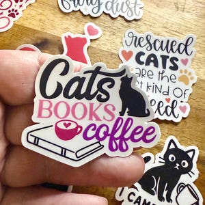 May include: A die-cut sticker featuring the text Cats in black, BOOKS in pink, and coffee in purple. The design includes a black cat silhouette, a white book, and a pink coffee cup with a heart in the foam. The sticker has a white border.