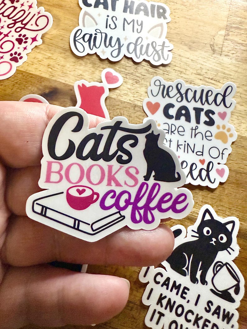 May include: A die-cut sticker featuring the text Cats in black, BOOKS in pink, and coffee in purple. The design includes a black cat silhouette, a white book, and a pink coffee cup with a heart in the foam. The sticker has a white border.