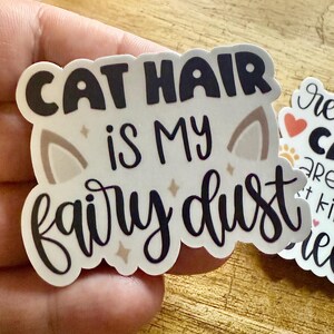 May include: A die-cut sticker with a white background featuring the black text CAT HAIR IS MY fairy dust. The design includes two light brown cat ear shapes and small four-pointed star accents in a light tan color.