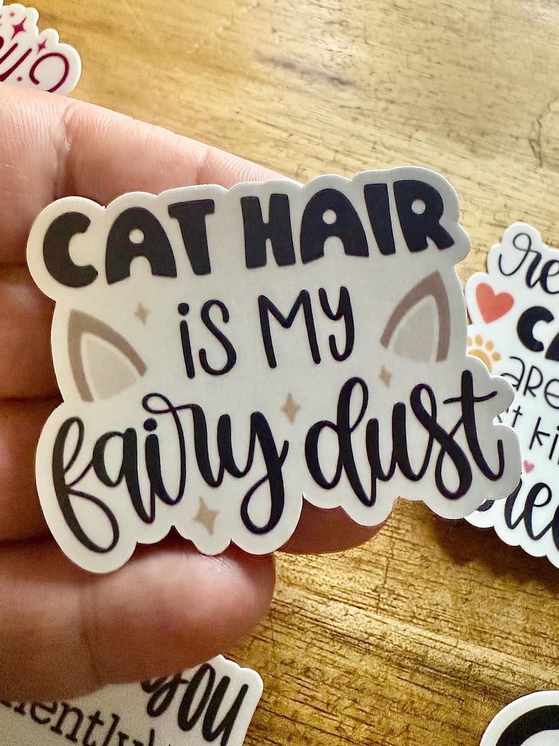 May include: A die-cut sticker with a white background featuring the black text CAT HAIR IS MY fairy dust. The design includes two light brown cat ear shapes and small four-pointed star accents in a light tan color.