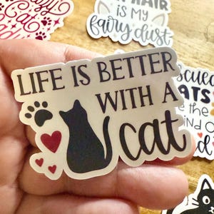 May include: A die-cut vinyl sticker featuring the black text "LIFE IS BETTER WITH A cat" alongside a black silhouette of a sitting cat, a black paw print, and three small red hearts. The sticker has a white border and a matte finish.