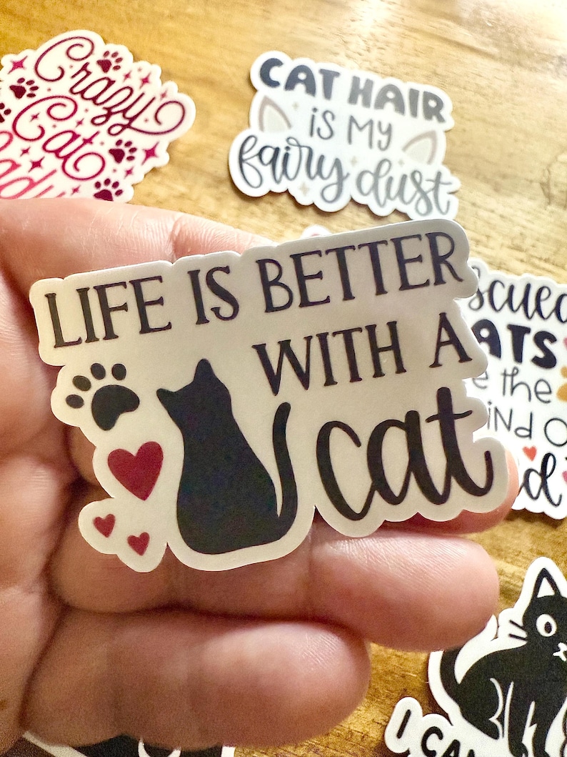 May include: A die-cut vinyl sticker featuring the black text "LIFE IS BETTER WITH A cat" alongside a black silhouette of a sitting cat, a black paw print, and three small red hearts. The sticker has a white border and a matte finish.