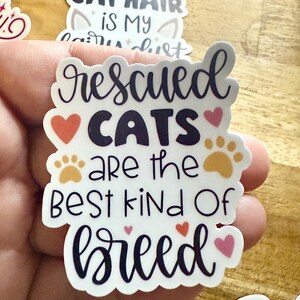 May include: A white die-cut sticker featuring the dark purple text 'rescued CATS are the BEST kind Of breed'. The design includes two orange paw prints, two red hearts, and two pink hearts arranged around the lettering on a white background.