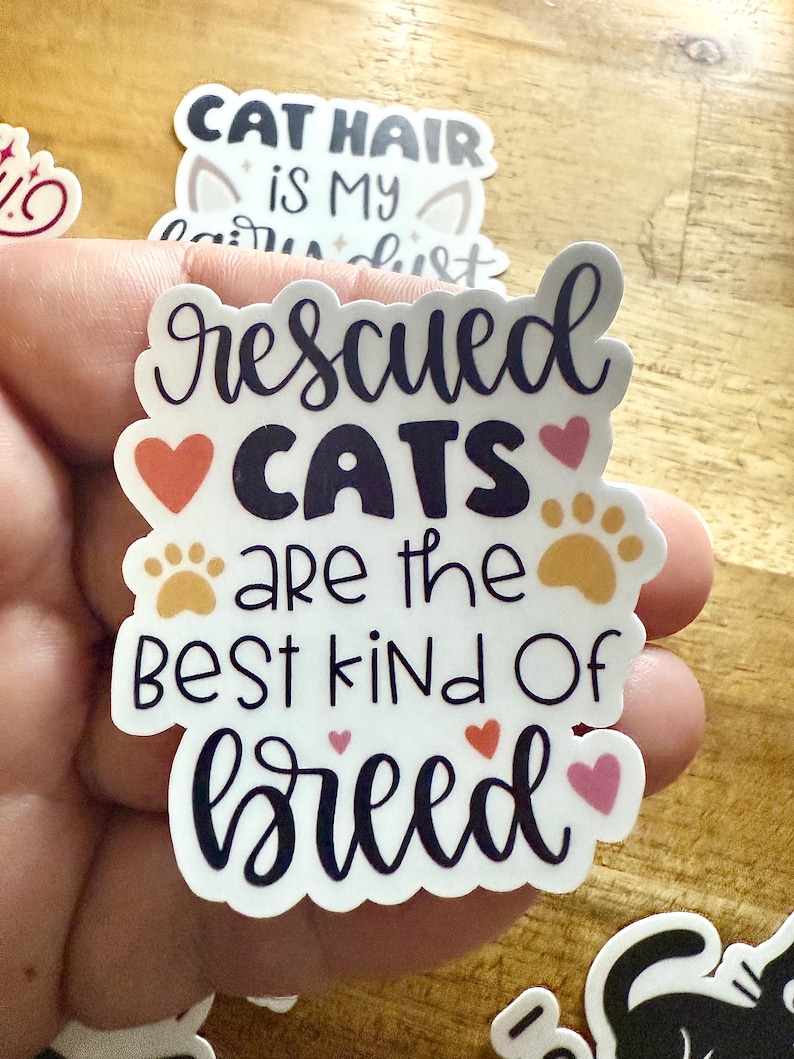 May include: A white die-cut sticker featuring the dark purple text 'rescued CATS are the BEST kind Of breed'. The design includes two orange paw prints, two red hearts, and two pink hearts arranged around the lettering on a white background.