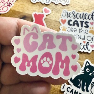 May include: A die-cut sticker featuring the text CAT MOM in a wavy, pink font. The letter O in MOM is replaced by a white paw print. The design is surrounded by small pink hearts and two small cat ears at the top, all on a white background.