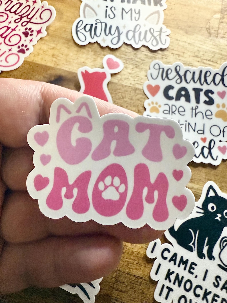 May include: A die-cut sticker featuring the text CAT MOM in a wavy, pink font. The letter O in MOM is replaced by a white paw print. The design is surrounded by small pink hearts and two small cat ears at the top, all on a white background.