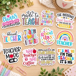 May include: A collection of twelve colorful vinyl stickers featuring teacher-themed phrases and illustrations, including apples, pencils, rainbows, and hearts, arranged on a light wood surface with office supplies nearby.