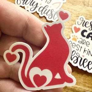 May include: A die-cut sticker featuring a solid red silhouette of a sitting cat. The cat has a heart-shaped cutout on its side, a heart at the tip of its tail, and a small pink heart floating above its head. The sticker has a white border.