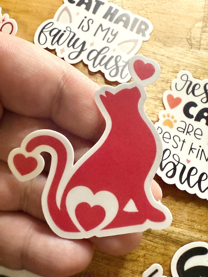 May include: A die-cut sticker featuring a solid red silhouette of a sitting cat. The cat has a heart-shaped cutout on its side, a heart at the tip of its tail, and a small pink heart floating above its head. The sticker has a white border.