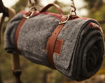 May include: A gray wool blanket with red and white stripes is rolled up and secured with brown leather straps. The blanket is hanging from two brown leather straps with metal clasps.