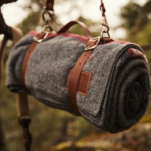 May include: A gray wool blanket with red and white stripes is rolled up and secured with brown leather straps. The blanket is hanging from two brown leather straps with metal clasps.