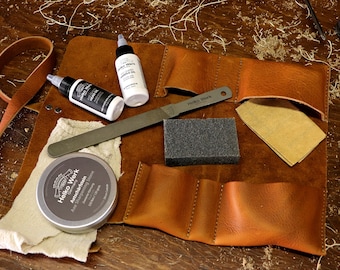 Puede incluir: Un kit de herramientas enrollable de cuero marrón con una correa, que contiene una lima de metal, un bloque de lijado gris, una pequeña lata de pasta de afilar y dos botellas de productos para el cuidado del cuero. El kit está etiquetado como "Helko Work".