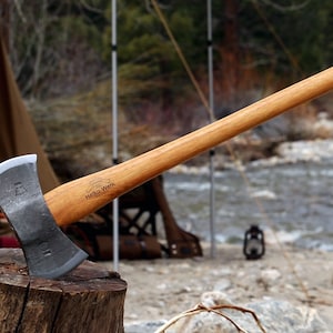 May include: A double-headed axe with a wooden handle. The axe head is made of steel and has a black finish. The handle is made of wood and has a natural finish. The axe is sitting on a wooden stump. The text "Helko Werk" is engraved on the handle.