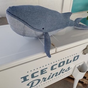 May include: A blue denim whale plush toy sitting on top of a white cooler with the text "ICE COLD DRINKS" printed on the side.