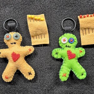 May include: Two handmade felt voodoo doll keychains, one yellow and one green, each with button eyes and a stitched heart. The yellow doll has black stitches, while the green doll has red stitches. Also shown are two felt pin holders.
