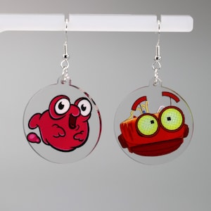 May include: A pair of clear acrylic earrings with red and green cartoon characters. The left earring features a red cartoon character with large eyes and a wide mouth. The right earring features a red robot with large green eyes.