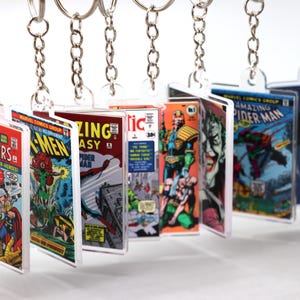 May include: A collection of comic book cover keychains featuring iconic Marvel and DC Comics characters. The keychains showcase vibrant artwork from titles like Avengers, X-Men, Spider-Man, and Batman. Each keychain is rectangular and attached to a silver chain.